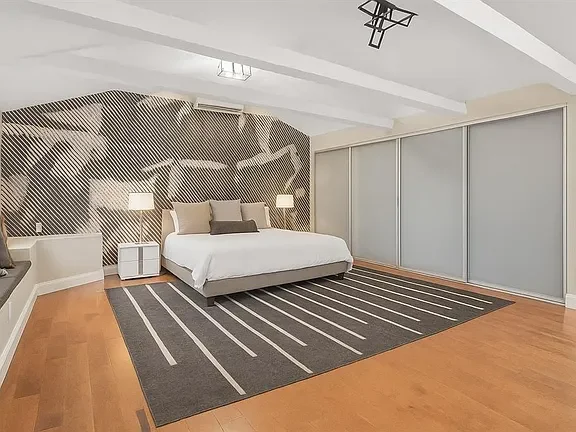 Modern bedroom featuring a large bed with white bedding, nightstands with lamps, a geometric accent wall, a striped area rug, and a large sliding closet door.