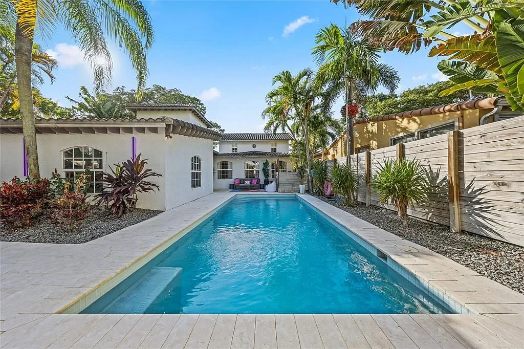 Backyard with a rectangular swimming pool, surrounded by tropical plants and palm trees, with a patio and seating area in the background under a clear blue sky.