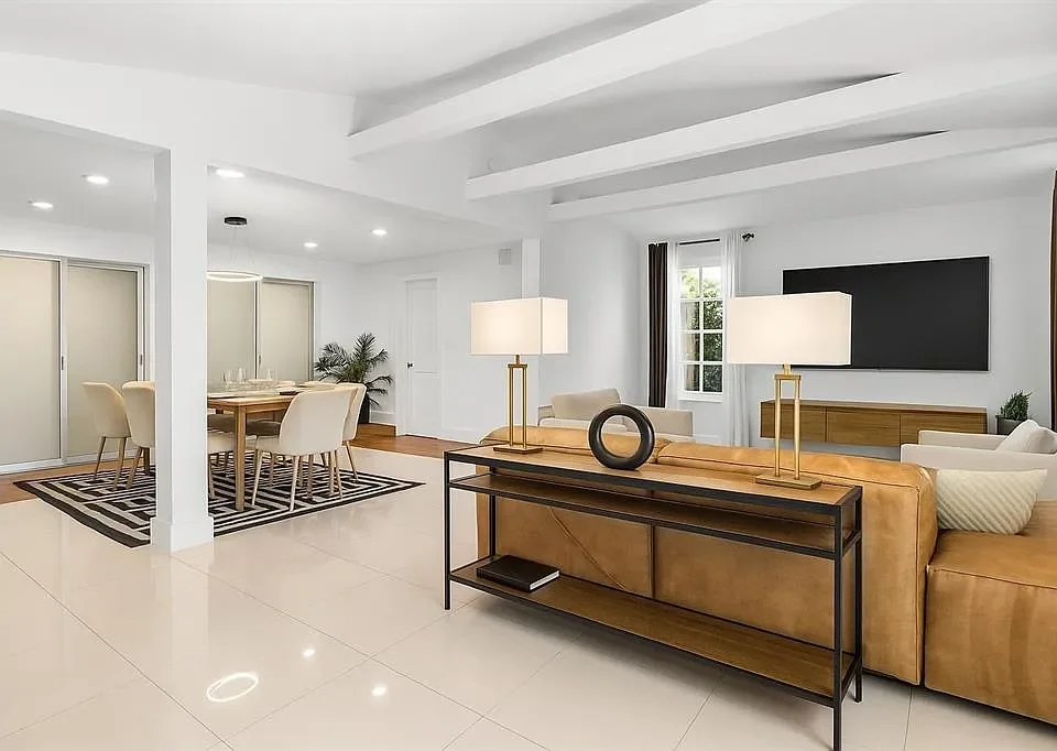 Modern, minimalistic living room with white walls, a beige sectional sofa, a wooden TV stand with a flat-screen TV, a black framed console table with lamps, and a dining area with a wooden table and cream chairs.