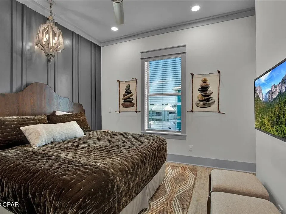 A bedroom with a wooden bed, brown textured bedding, and a white pillow. There is a hanging lamp on the wall, decorative curtains with rock art, and a window showing buildings outside. A wall-mounted TV is visible on the right, with a beige bench below it, and a patterned rug on the floor.