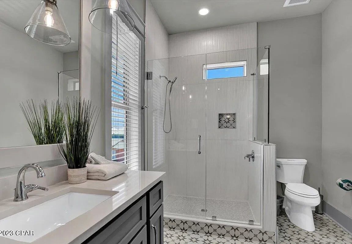 Modern bathroom with gray cabinets, large mirror, plant, towel, shower with glass doors, small window, and toilet.