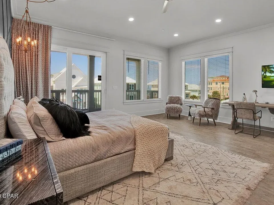 A spacious bedroom with a large bed, multiple pillows, and a cozy blanket. There is a nightstand with books and a lamp. The room has large windows with blinds and views of neighboring houses. Two plush chairs and a small desk are arranged near the wi