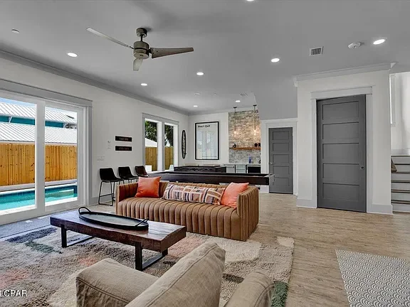 Living room with beige and striped sofas, a wooden coffee table, a large rug, and a ceiling fan, opening to a backyard with a pool through sliding glass doors.