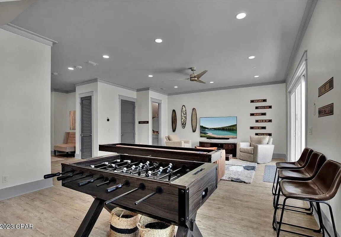 Living room with a foosball table, a large flat-screen TV, two white armchairs, a beige sofa, and three barstools against a wall. There are decorative wall signs and a window with white curtains.