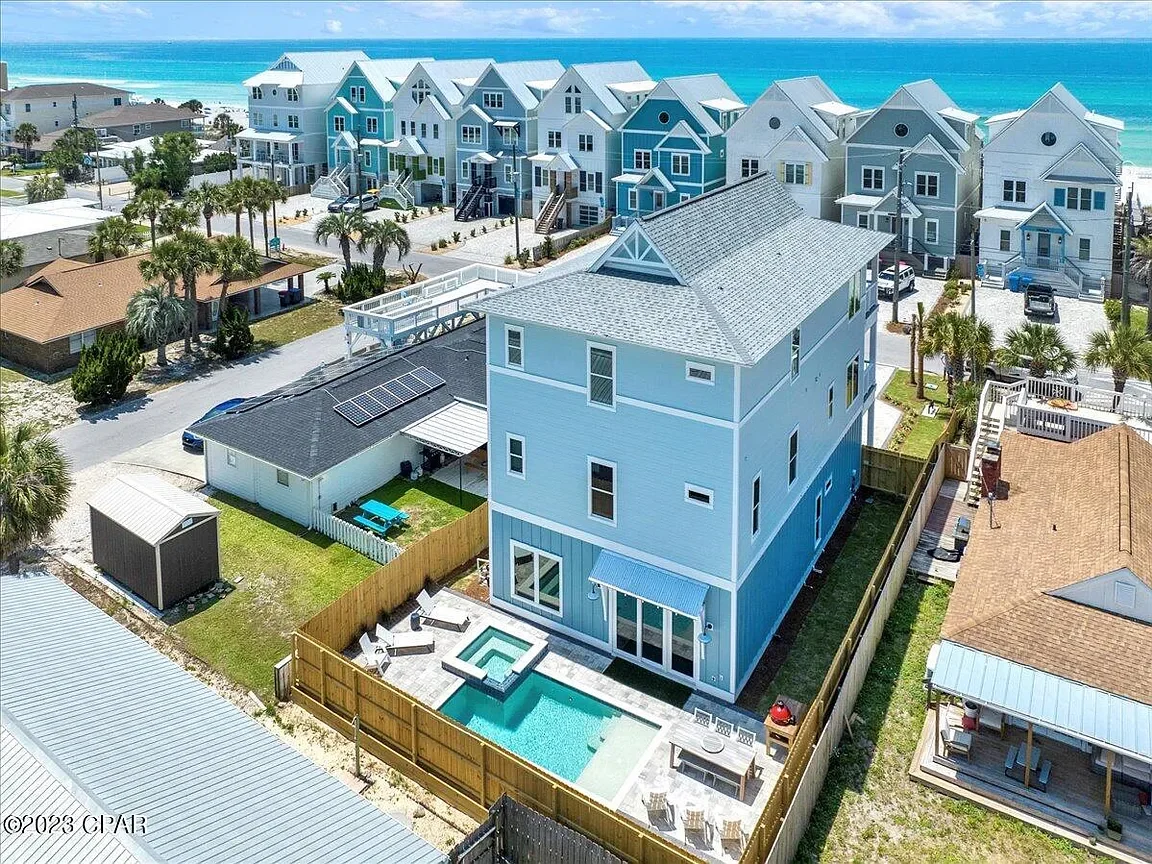 An aerial view of a beachside neighborhood with colorful houses, including a large blue house with a fenced backyard, swimming pool, and hot tub, located near the ocean with sandy shores and waves in the background.