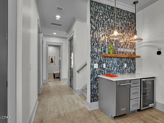 Interior view of a modern home with white walls, hardwood flooring, a hallway leading to other rooms, and a kitchen area with a mosaic tile accent wall, pendant lights, a wooden shelf, and a gray countertop with cabinets.