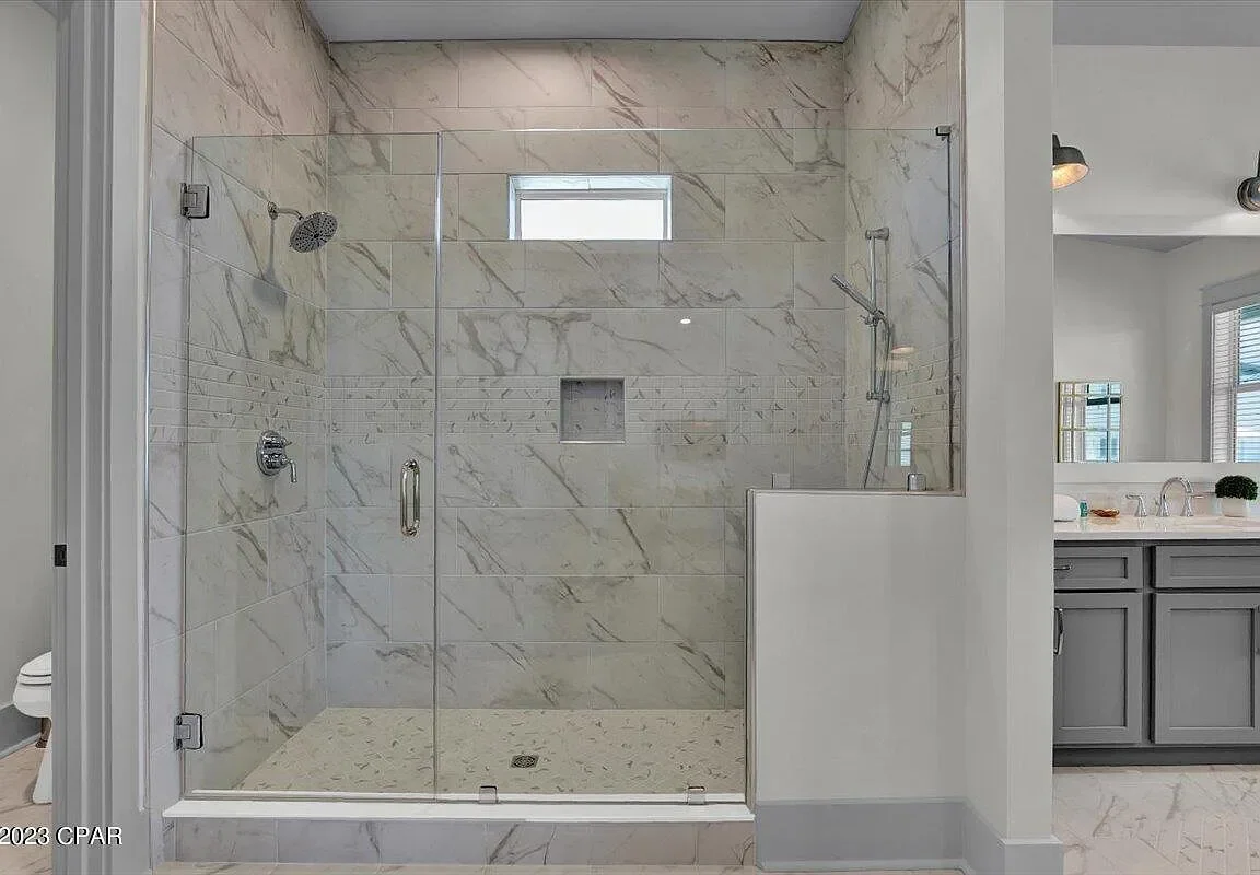 A modern walk-in shower with glass doors and marble tile walls and floor, located in a bathroom with a gray vanity and two sinks visible in the background.