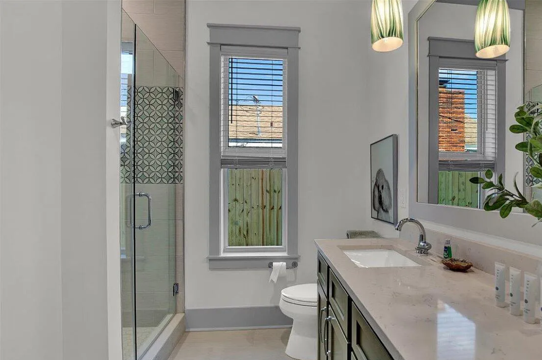 Modern bathroom with a glass-enclosed shower, double window with blinds, white walls, gray trim, a vanity with a single sink, large mirror, and green pendant light fixtures.