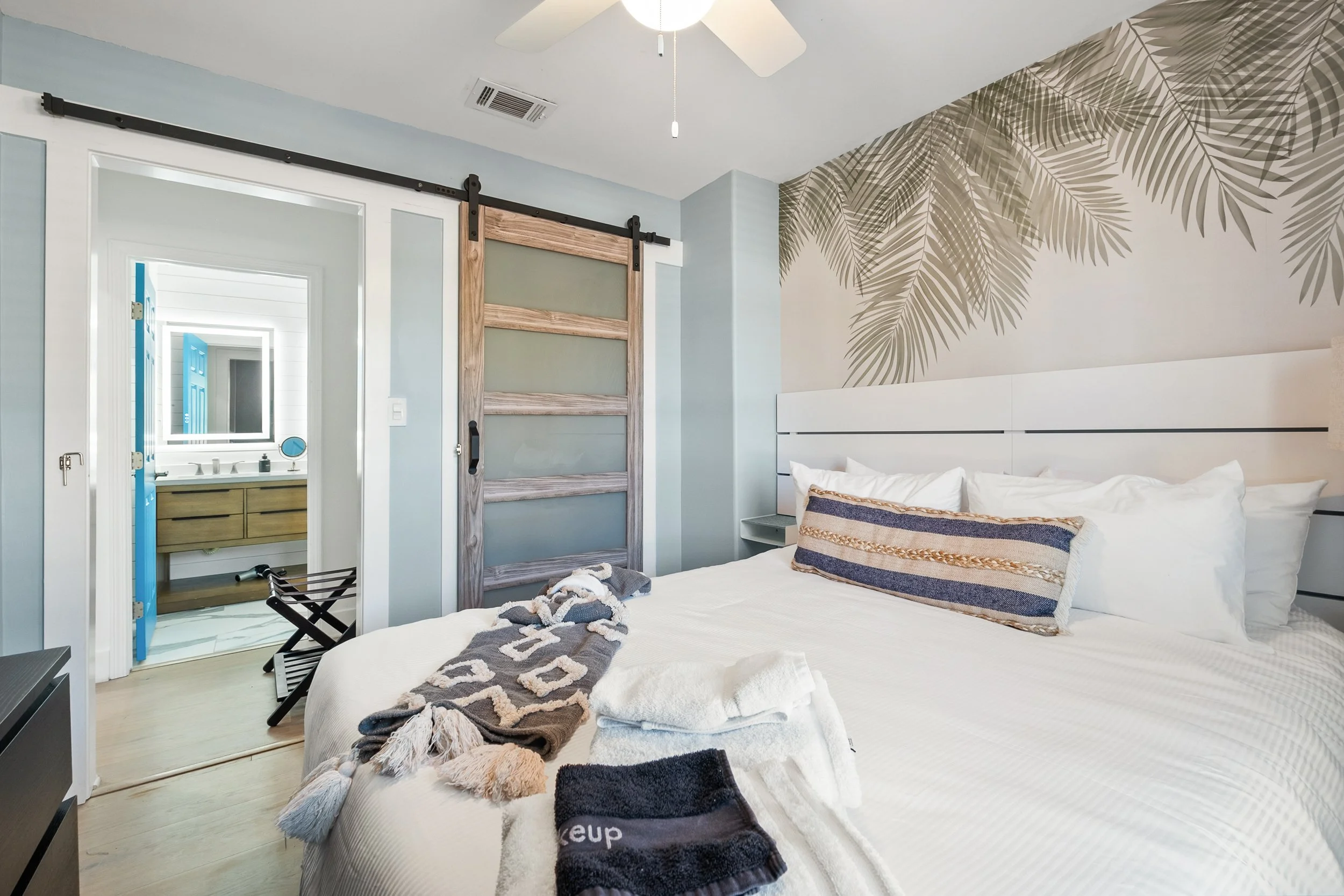 A bedroom with a large bed covered in white linens, a decorative pillow, and towels on top, with a wallpapered wall featuring leaf patterns. A sliding barn door is open to a bathroom with a blue door and vanity, and a laundry basket is on the floor.