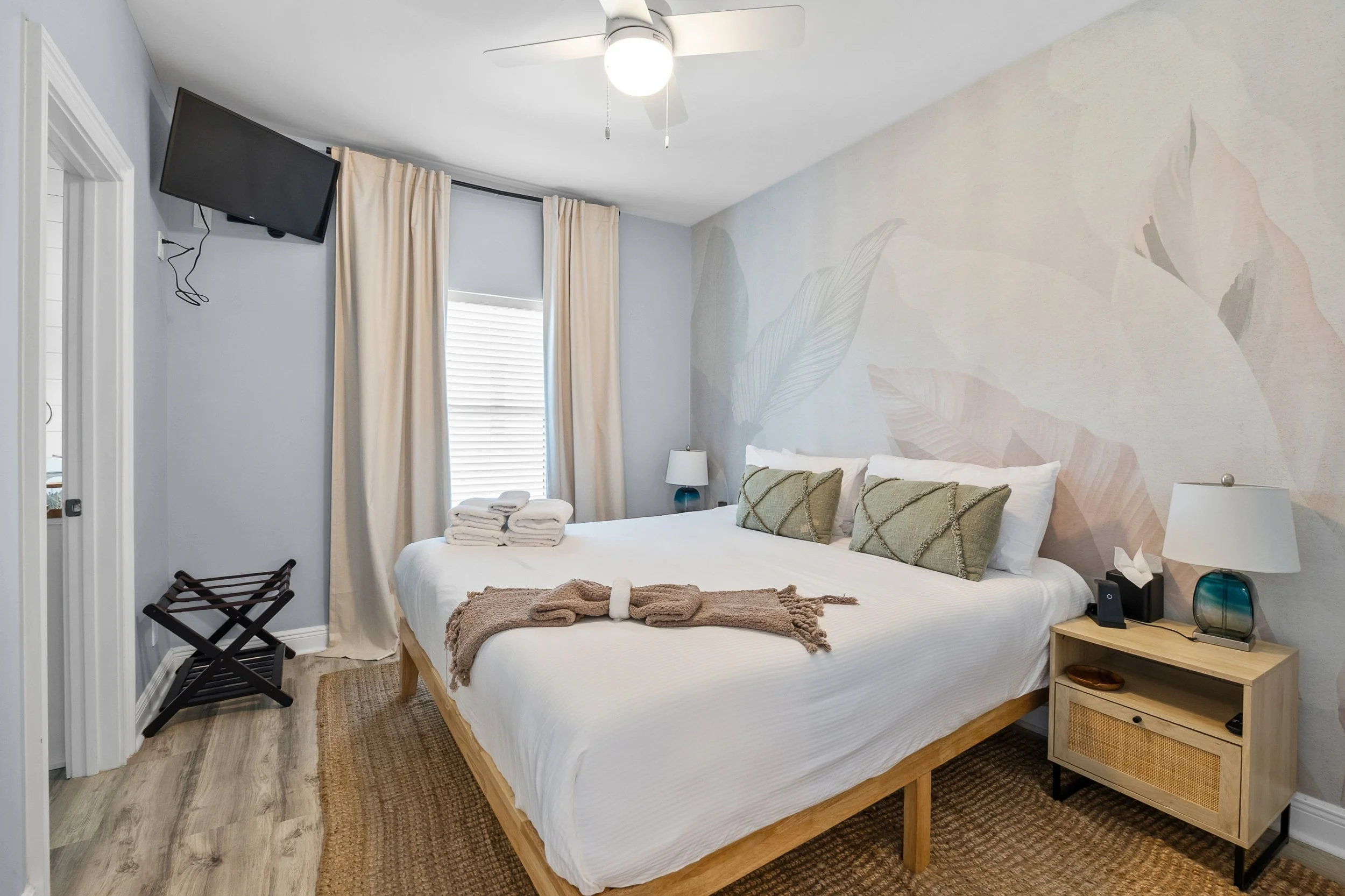 A bedroom with a large bed, pillows, and blankets, a nightstand with a lamp and tissue box, a wall with a floral mural, a window with curtains, a ceiling fan, and a small TV mounted on the wall.