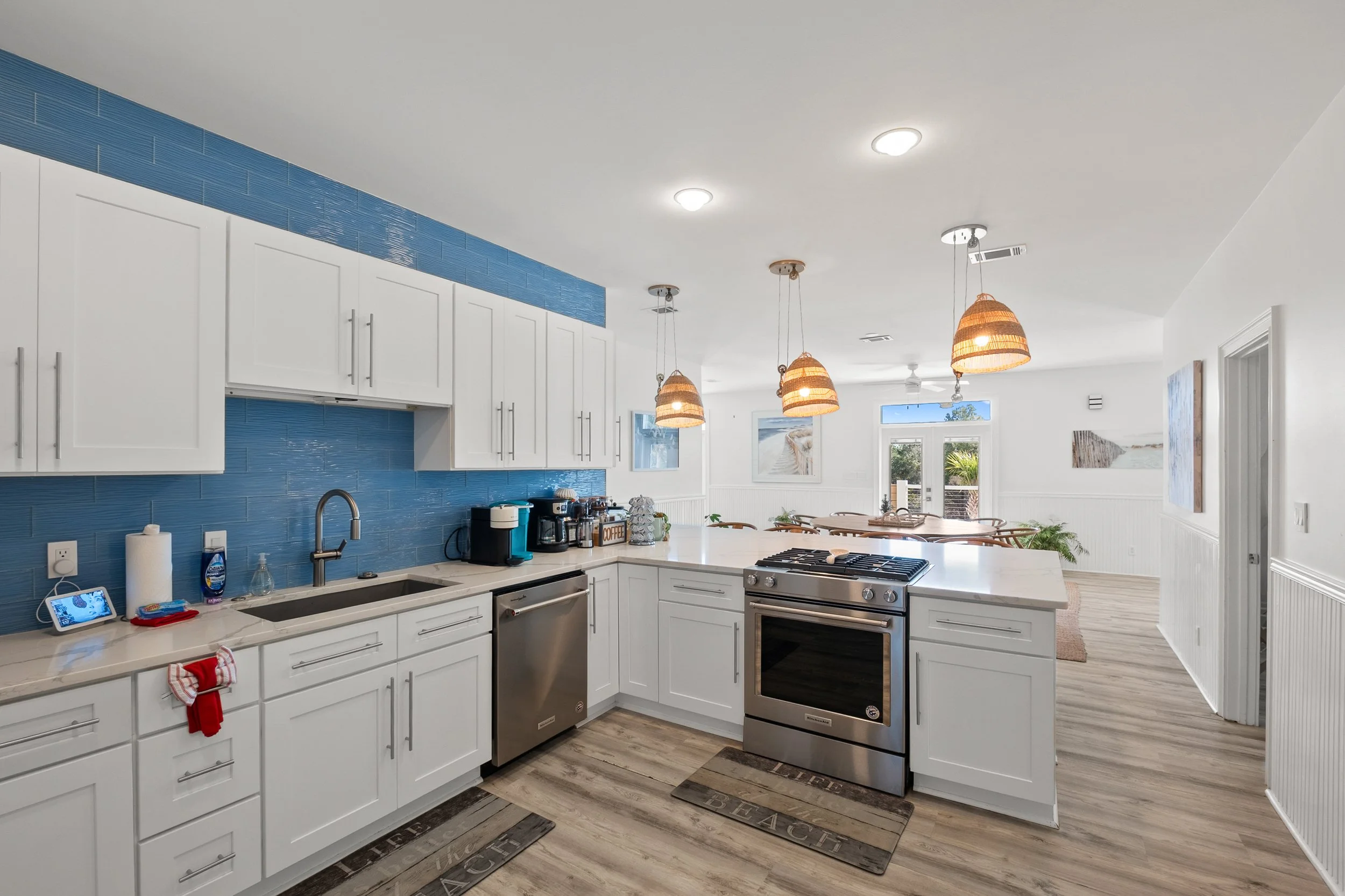 Modern kitchen with white cabinets, stainless steel appliances, and a blue tiled backsplash, with a view of a dining area and patio outside.