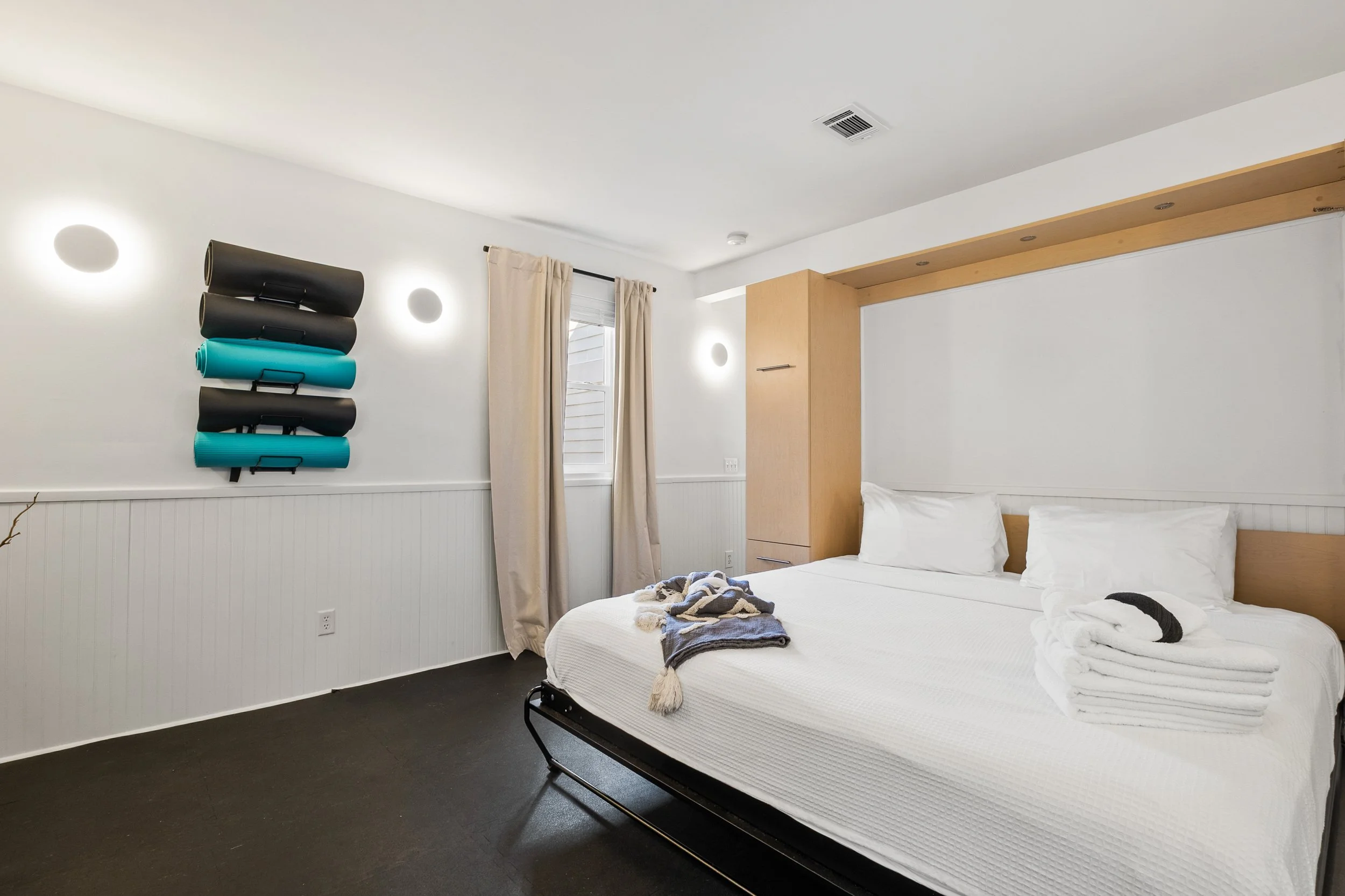 A modern bedroom with a neatly made bed, a small wooden wardrobe, beige curtains, a window, and wall-mounted yoga mats.