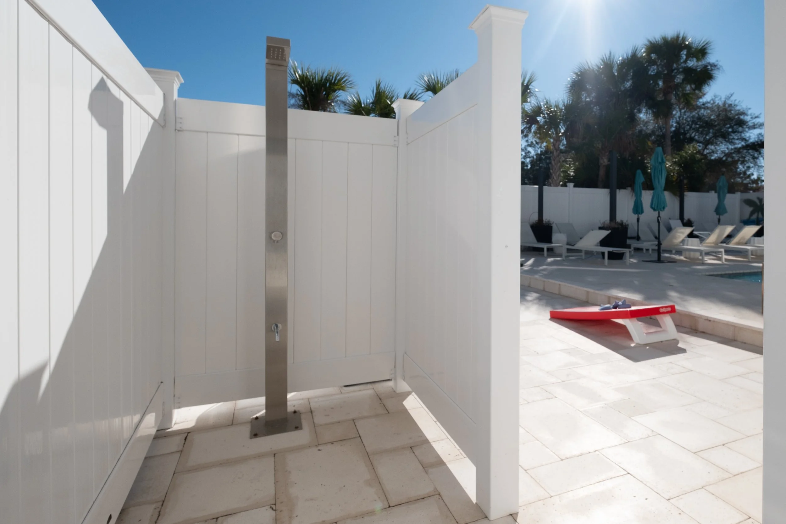Outdoor pool area with lounge chairs, umbrellas, and palm trees in the background under a sunny sky, with a white fence surrounding the space.