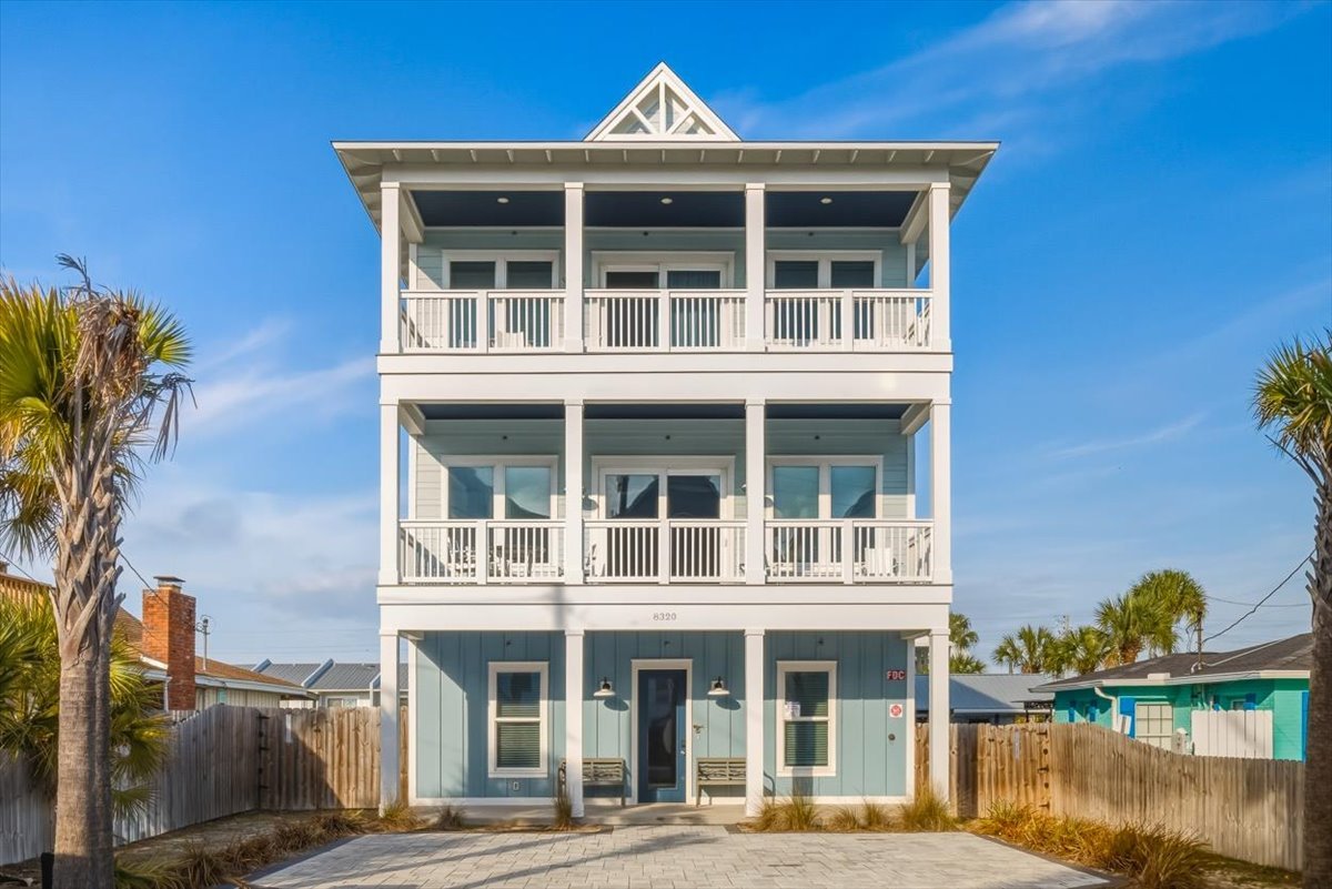 A three-story beach house painted in light blue with white trim, featuring balconies on each level, surrounded by palm trees and a paved walkway in the front.