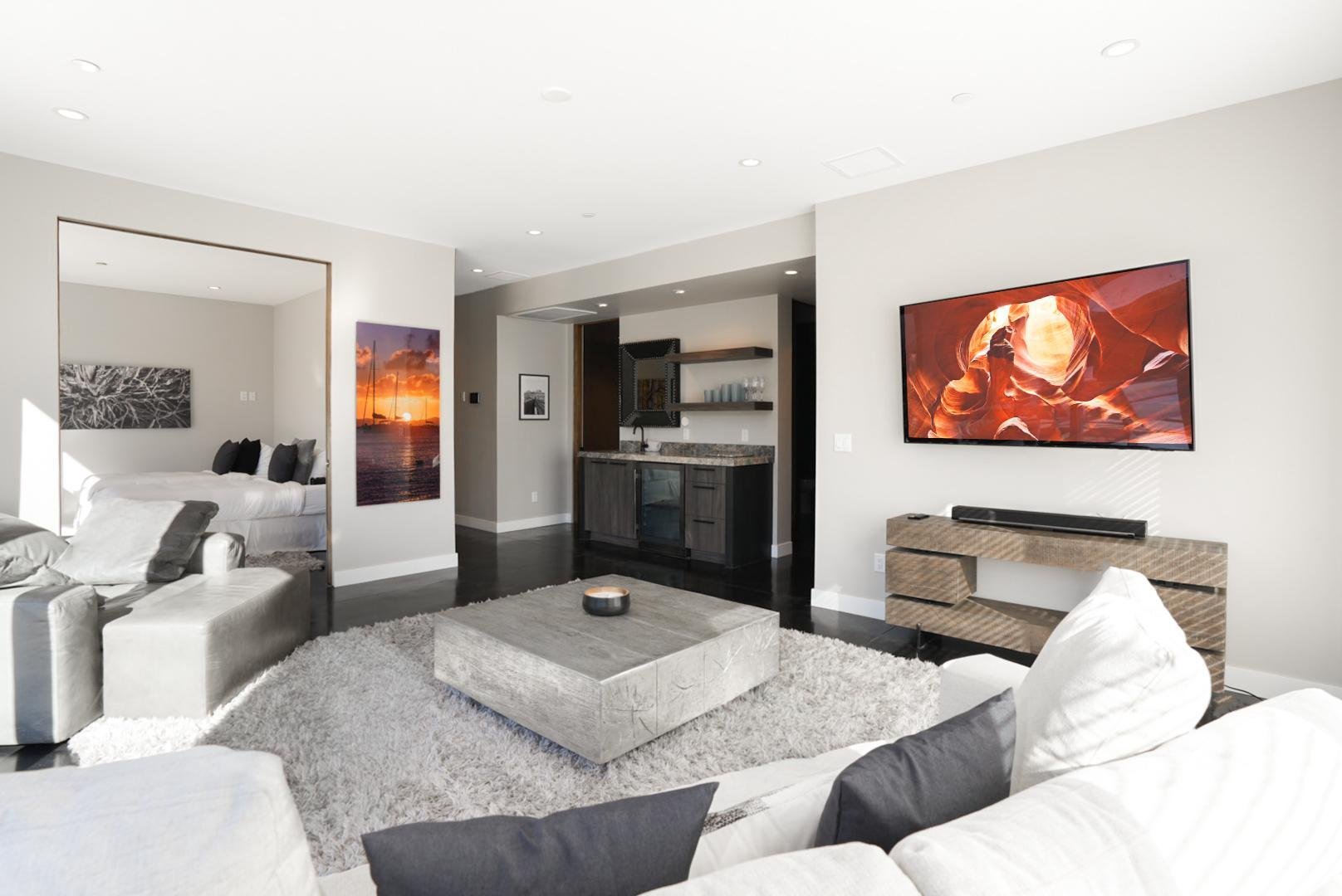 Modern living room with a large white sofa, a matching armchair, and a gray area rug. A framed picture of a sunset over the water hangs on the wall. A flat-screen TV displays a canyon scene. A small kitchen area with bar shelves and countertop is vis