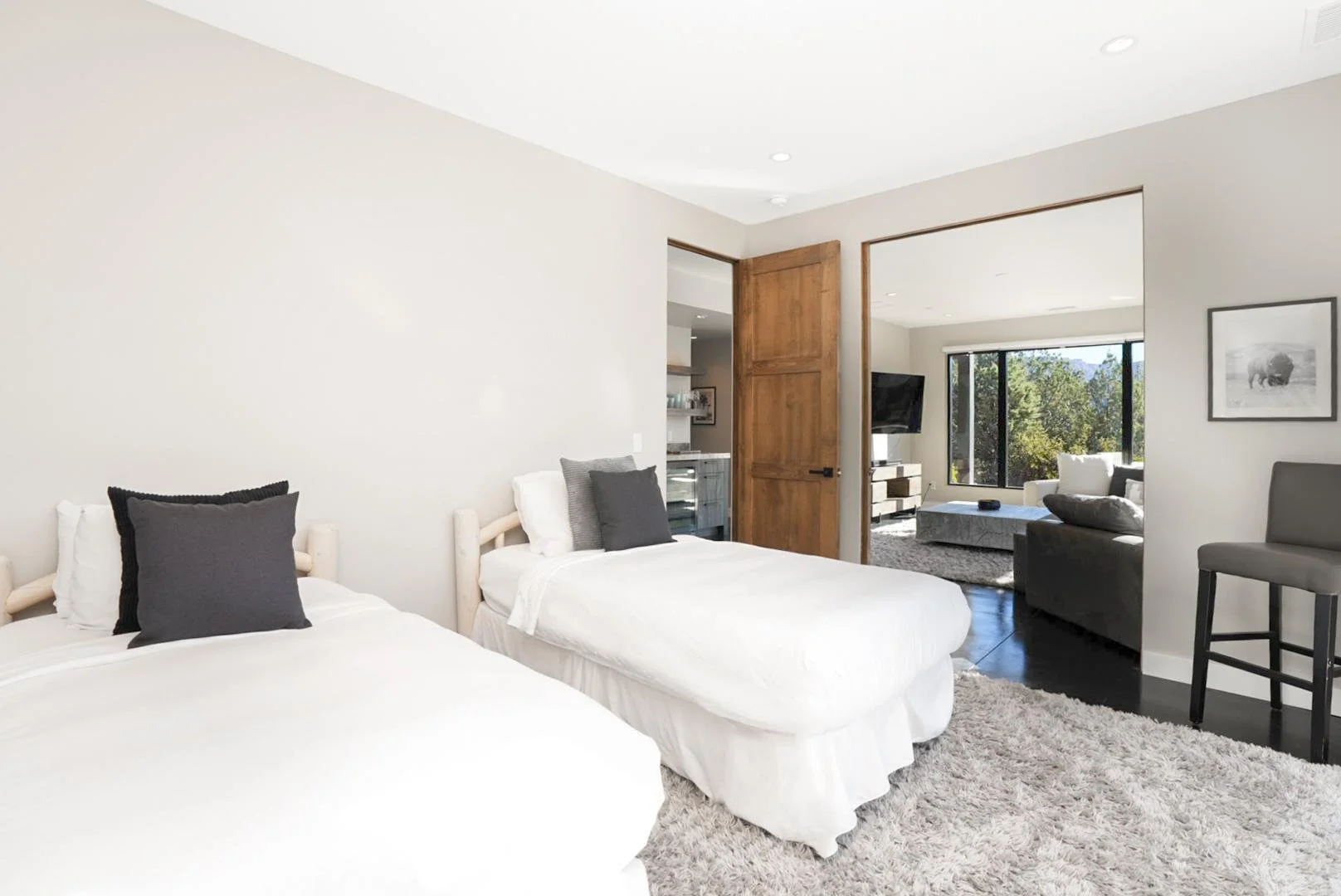 A living room with two beds and a seating area. The beds have white linens and dark pillows. The room features a wooden door, a shaggy rug, and a small open space leading to another seating area visible through an opening.