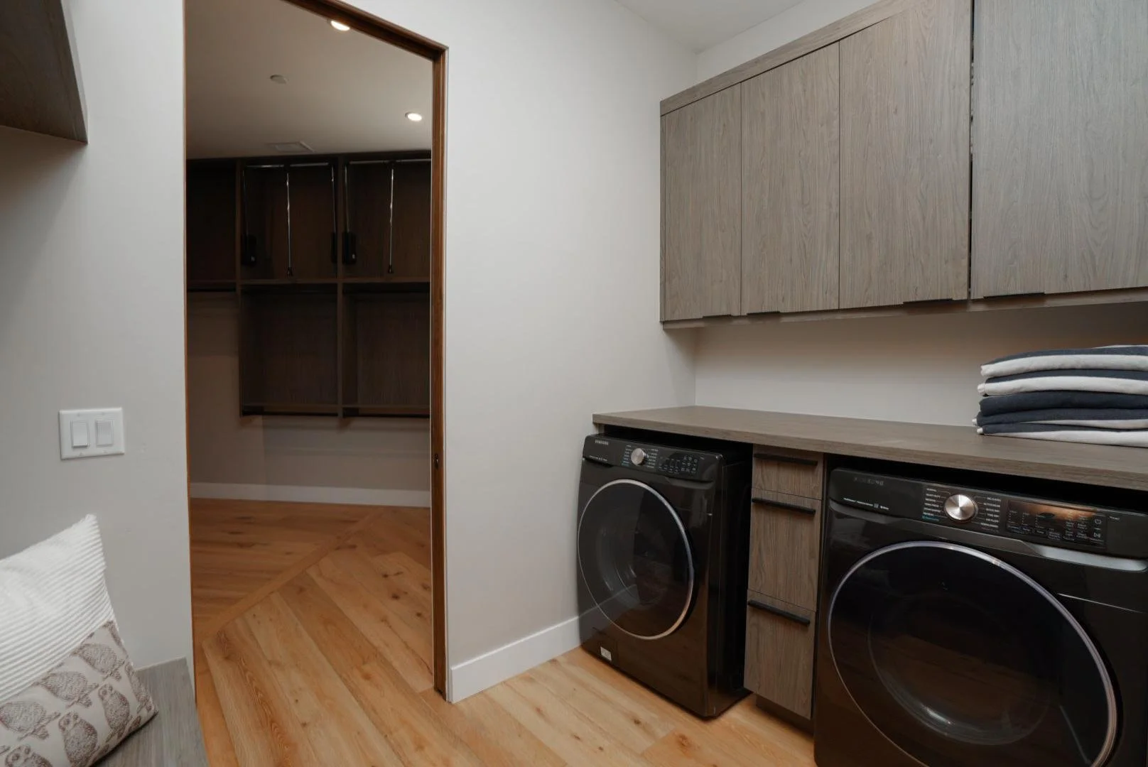 Laundry room with stacked washer and dryer, wooden cabinets, folded towels on countertop, tiled wooden floor, and plain white walls.