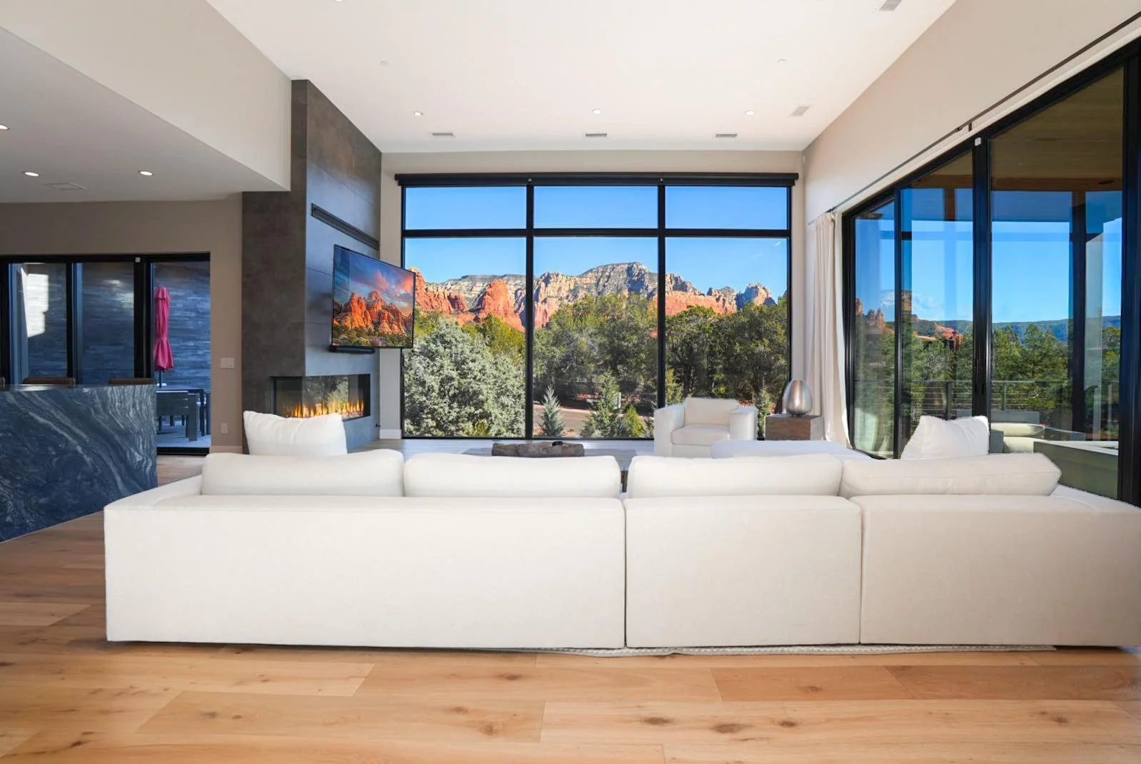 Modern living room with large windows showcasing a mountain landscape; white sofa and armchair, fireplace, flat-screen TV, and wooden flooring.