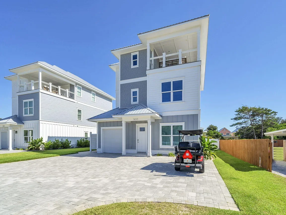 Modern three-story beach house with a driveway, golf cart, and a fenced yard.