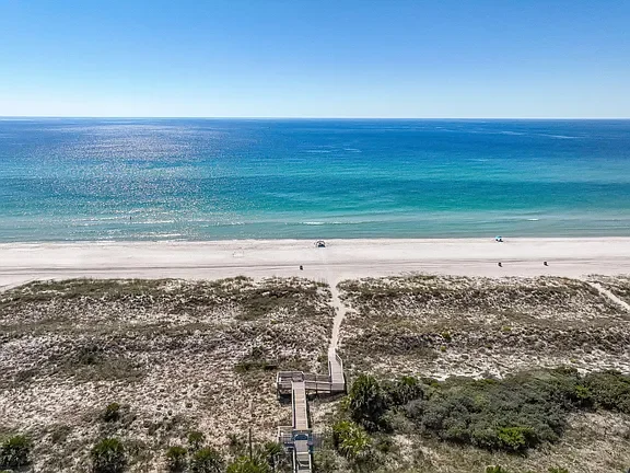 Aerial view of a beach with white sand, ocean waves, and a clear blue sky. A wooden boardwalk leads from the dunes to the beach.