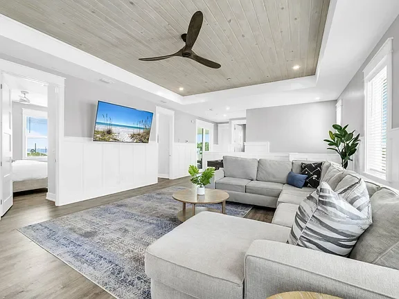 Bright living room with white walls, a gray sectional sofa with striped pillows, a wooden coffee table, a flat-screen TV on the wall, large windows with blinds, a gray rug, and a ceiling fan.