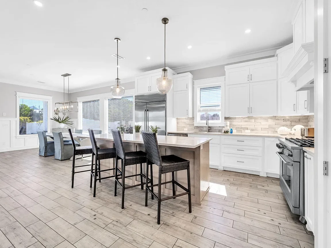 Bright, modern kitchen with white cabinets, a large island with four dark-colored chairs, stainless steel refrigerator, gas stove, light gray walls, and wooden flooring.