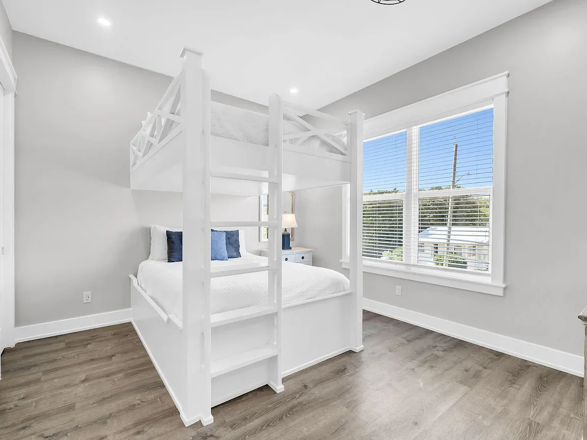 Bright bedroom with large window, white walls, hardwood floor, white bunk bed, and white nightstand with a lamp.