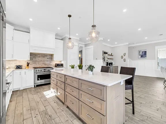 Modern kitchen with white cabinets, a large island with storage drawers, and pendant lighting, open to a living space with wood flooring.