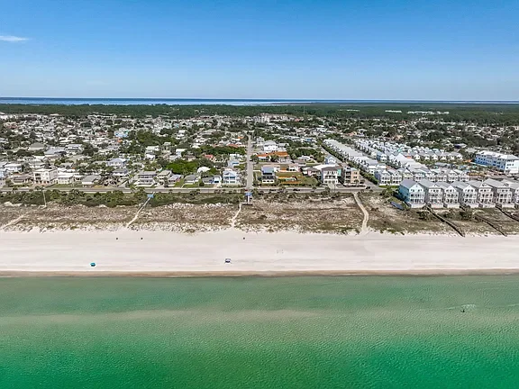 Aerial view of a beach town with houses and buildings near the shoreline, with the ocean in the background under a clear blue sky.
