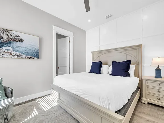Bedroom with white walls, a large bed with white bedding, two navy blue pillows, and a beige headboard. There's a nightstand with a blue lamp, a painting of a rocky coast, and natural light coming through a window.