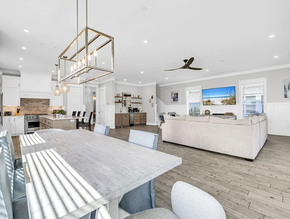 Open-concept living and dining area with white walls, hardwood floors, a large beige sectional sofa, wall-mounted TV, and a modern chandelier over the dining table.