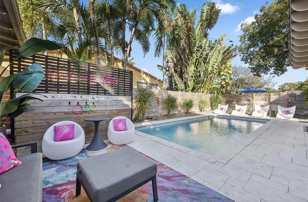 Backyard with swimming pool, tropical plants, lounge chairs with pillows, and a wooden privacy fence. Decor includes pink neon sign reading 'LOVE IS THE VIEW' and colorful hanging rings.