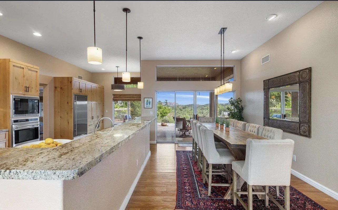 Open-concept kitchen and dining area with a granite countertop island, wooden cabinets, stainless steel appliances, and a dining table with cushioned chairs. Large sliding glass door leads to an outdoor space with a scenic view.