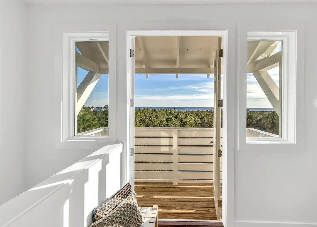 View from a balcony with white walls and railing, showing a landscape of trees and a distant ocean with a partly cloudy sky.