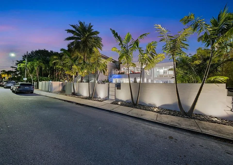 Modern house with white walls and large glass windows, surrounded by palm trees, at dusk with a purple and blue sky.