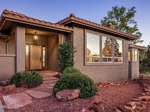 Front view of a beige stucco house with a brown tiled roof, large front windows, a wooden front door, and a landscaped yard with bushes, rocks, and a stone pathway under a clear sky.