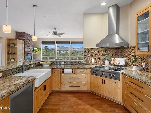 Modern kitchen with wooden cabinets and granite countertops, stainless steel range hood, pattern backsplash, ceramic sink, and large windows showing a scenic view of trees and hills outside.