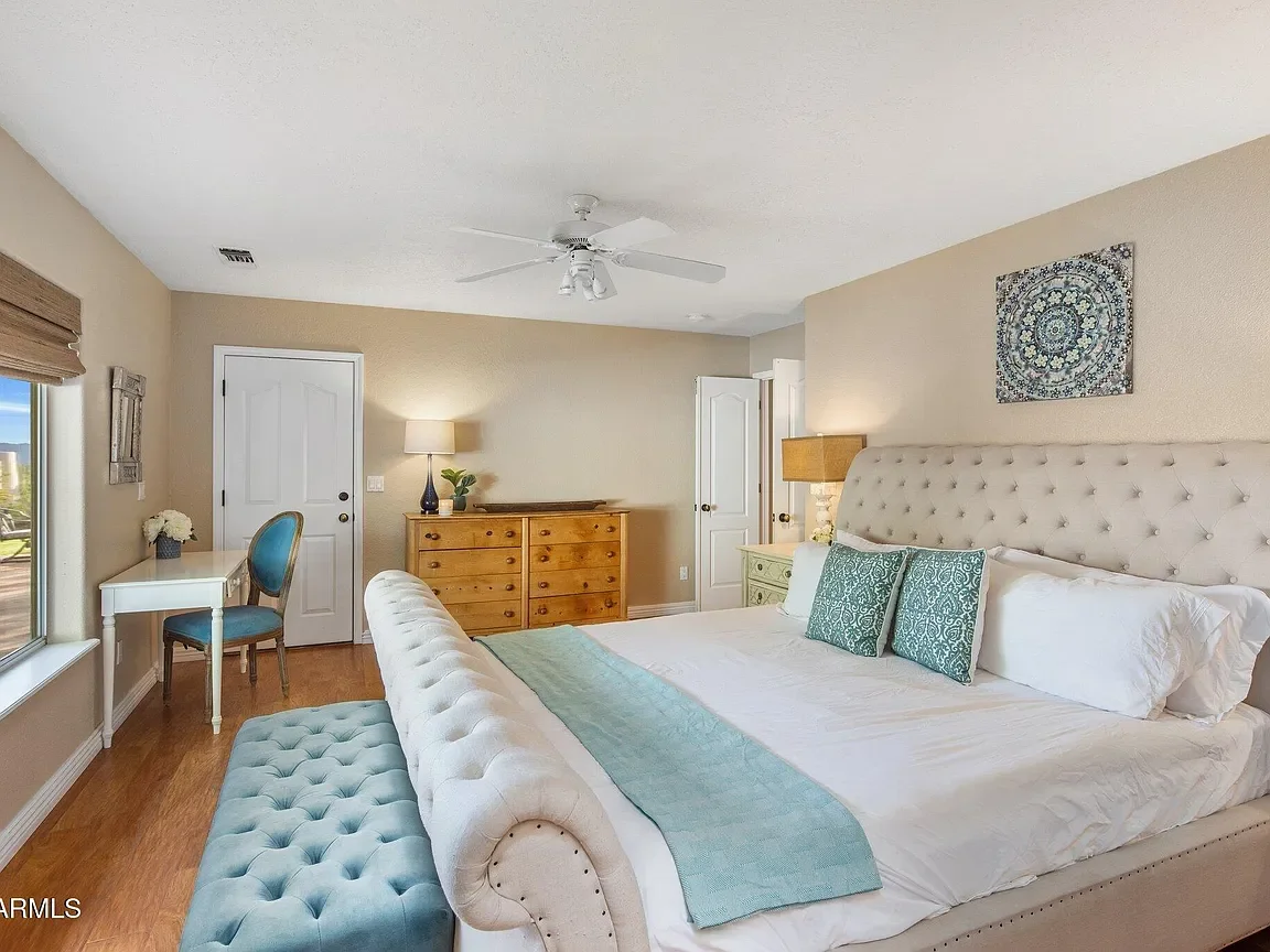 A bedroom with a large white tufted bed with two patterned teal pillows, a teal bed runner, and a beige upholstered bench at the foot of the bed. There are two nightstands with lamps, a wooden dresser, a small white writing desk with a chair, and a w