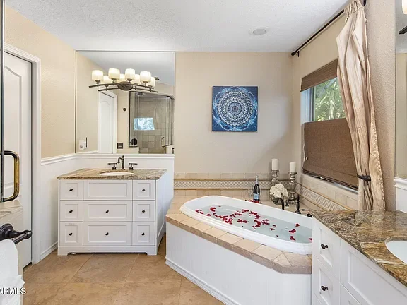 Bathroom featuring a bathtub with rose petals, white cabinetry, a granite countertop, a mirror, a shower area, and a window with curtains.