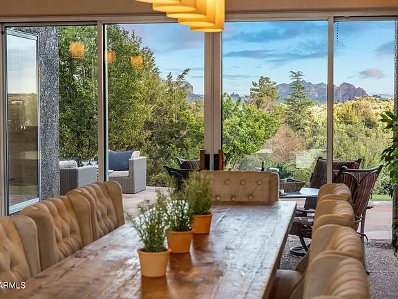 A dining room with a large wooden table, tan chairs, potted plants, and large glass windows overlooking a lush green outdoor landscape with trees and distant mountains.
