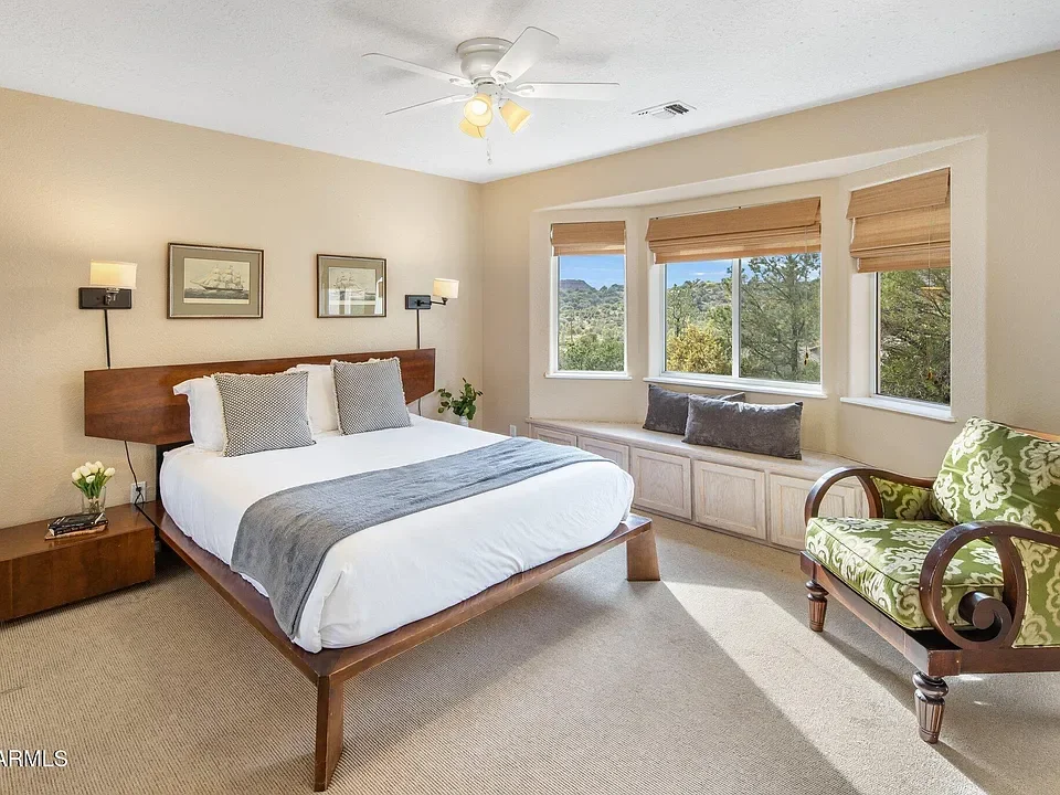 A bedroom with a wooden bed, white bedding, and gray pillows. There are three framed black and white pictures above the bed. A small wooden nightstand with a small pot of white flowers is on the left. The room has a bay window with three sections, be
