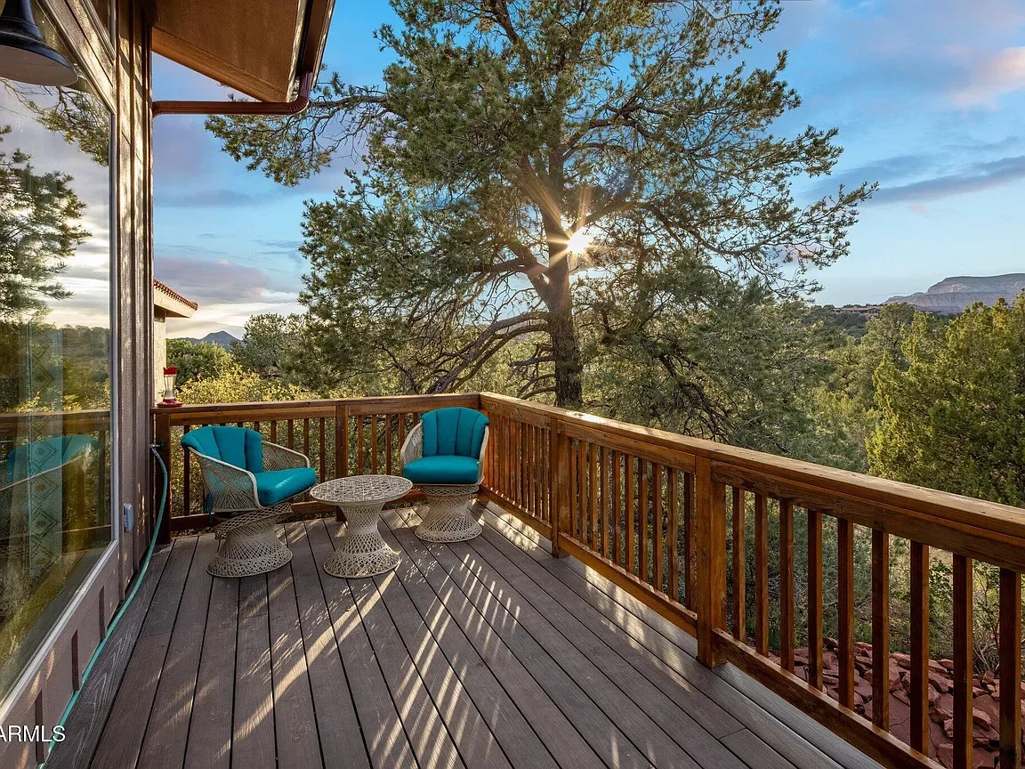 A wooden balcony with two blue cushioned chairs and a matching small round table, overlooking a green landscape with trees and mountains in the distance during sunset.
