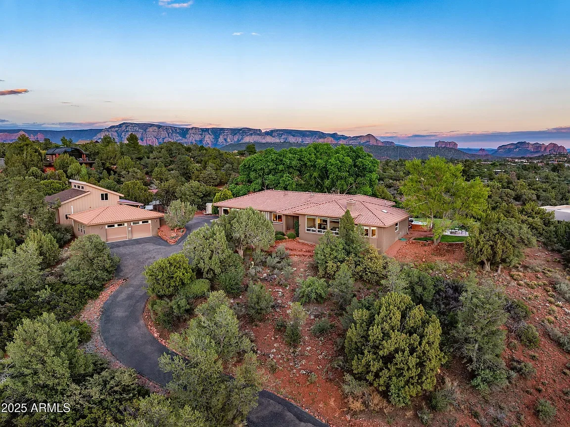 Aerial view of a house surrounded by trees and red desert landscape with mountains in the background during dusk.
