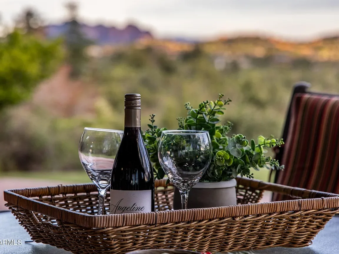 A wicker tray holding a wine bottle labeled "Angeline" and two empty wine glasses, with a potted green plant in the background, set outdoors on a sunny day.
