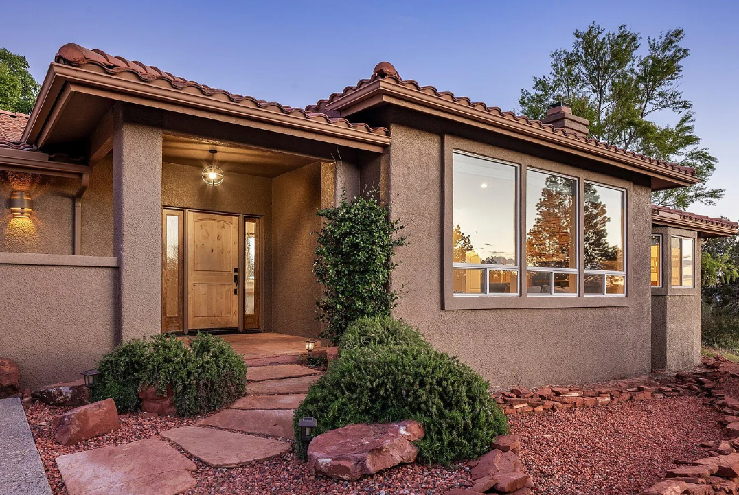 Front view of a modern house with a wooden front door, large windows, and a landscaped yard with bushes, rocks, and pathway at sunset.