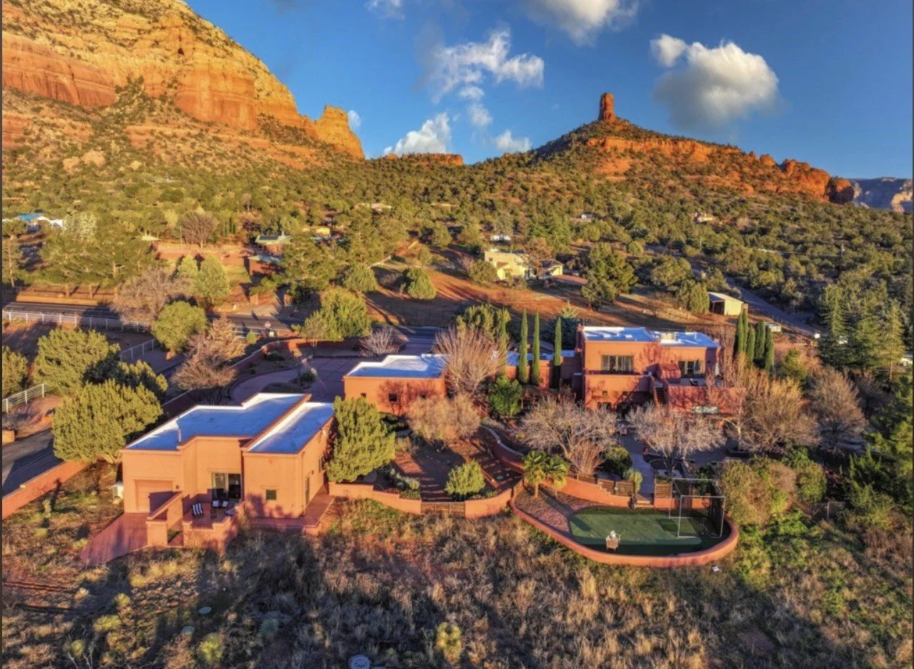 Aerial view of a residential area in Sedona, Arizona, with red rock formations in the background and several modern houses surrounded by trees, sunny sky, and a small sports court.
