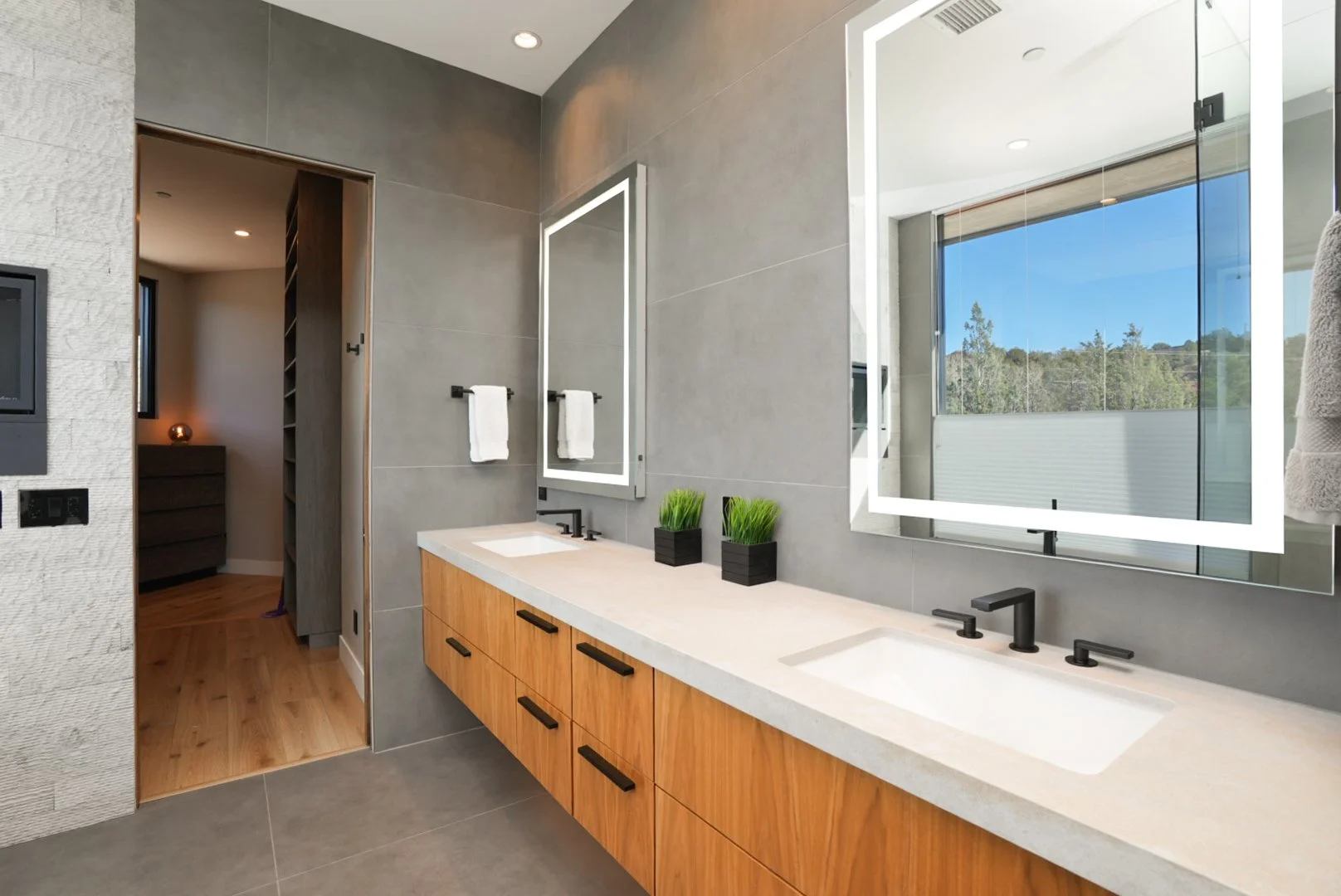 Modern bathroom with double sink vanity, large mirror with lighting, gray wall tiles, wooden cabinets, potted plants on vanity, and window showing outdoor scenery.