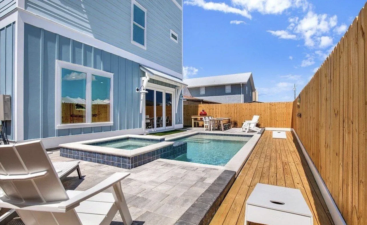 Backyard with a small swimming pool, hot tub, and wooden deck. There's a blue house with a sliding glass door, white trim, and a privacy fence. Patio furniture is visible, and the sky is blue with some clouds.