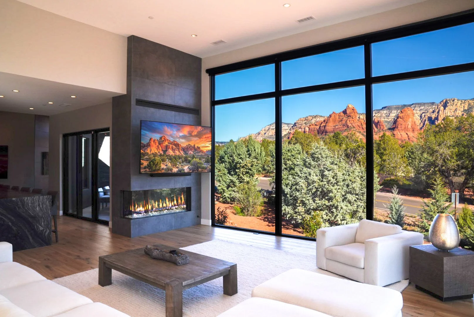 Modern living room with large floor-to-ceiling windows showing desert mountain scenery, fireplace, flat-screen TV, white sofa and armchair, wooden coffee table, and decorative vase.