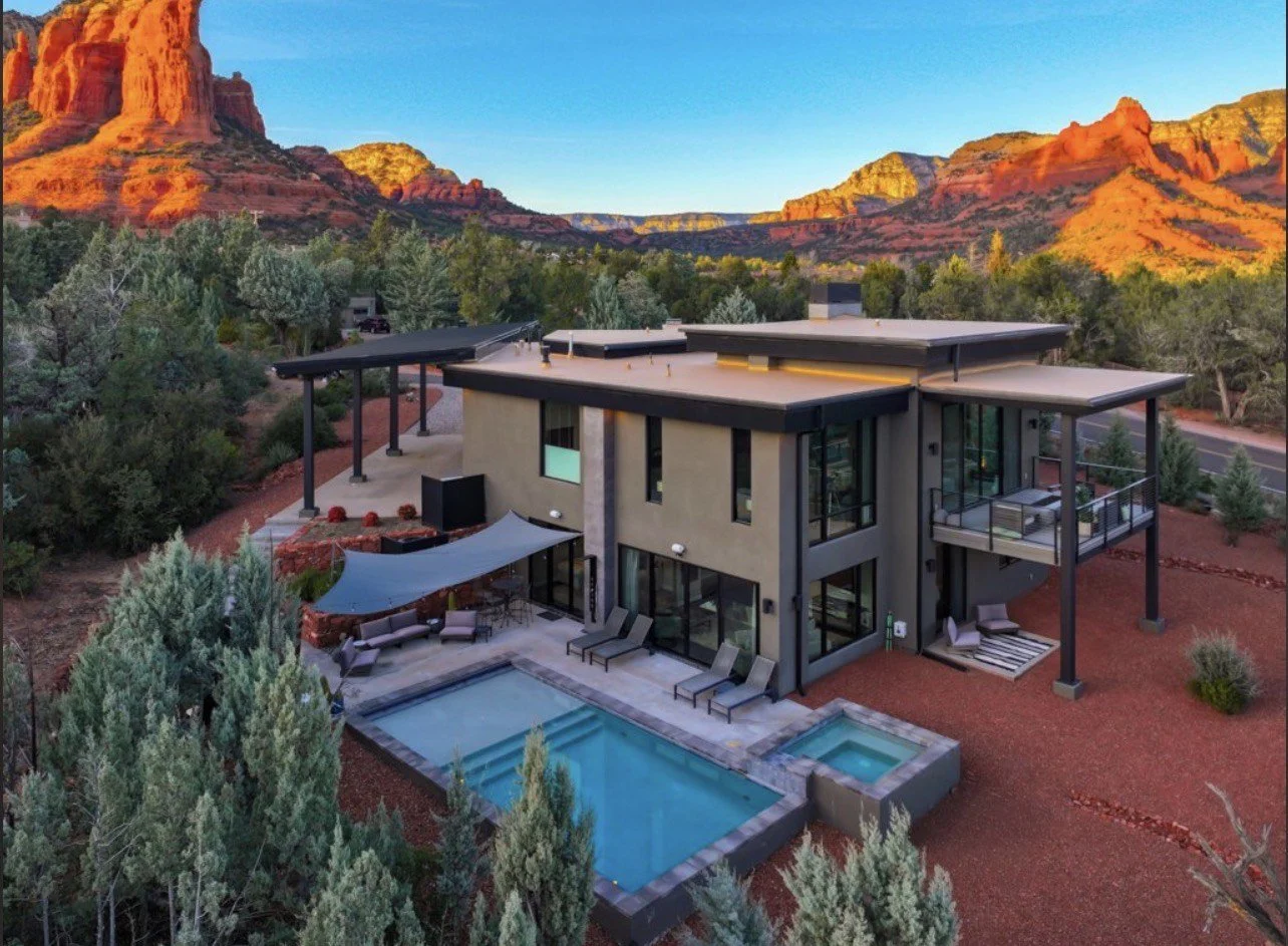 Modern house with outdoor pool and hot tub, surrounded by trees, with red rock mountains in the background during sunset.